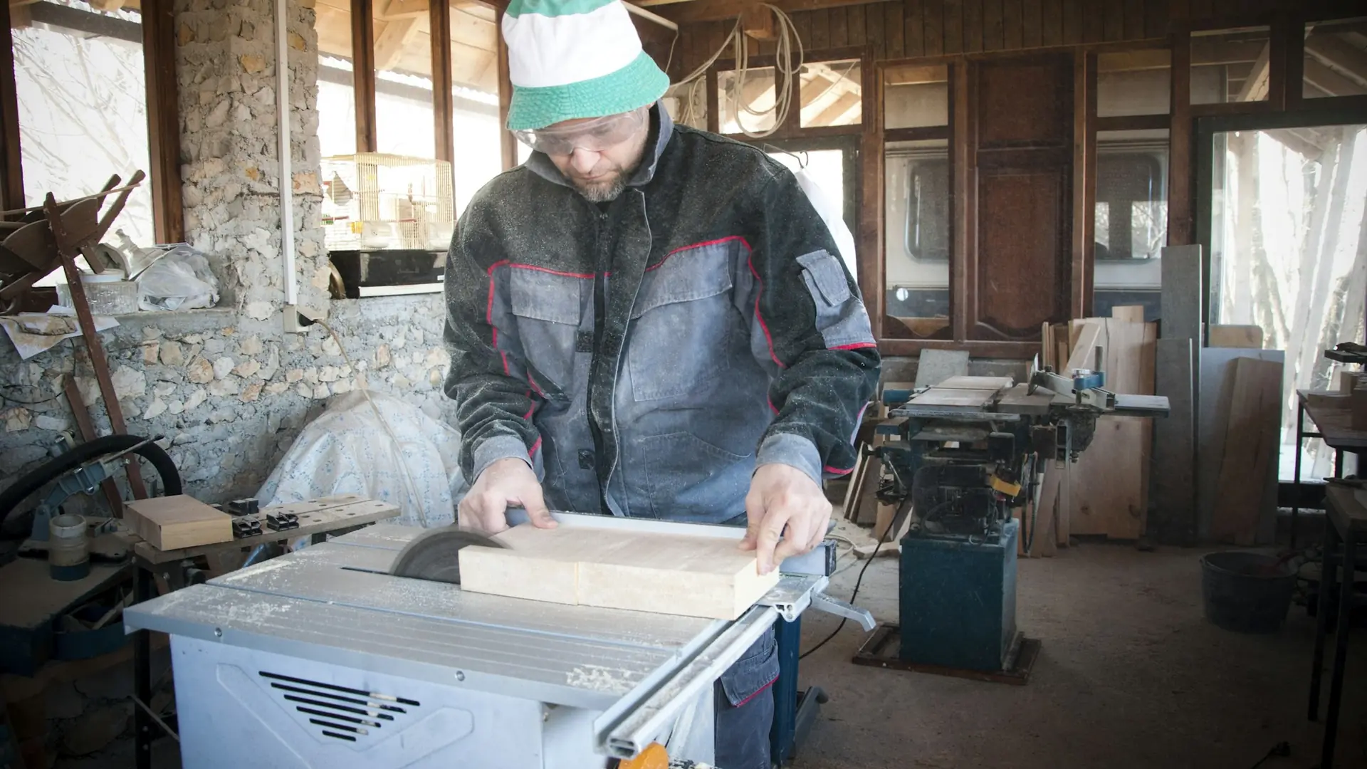 A woodworker in safety glasses feeding a board across a table saw in a workshop